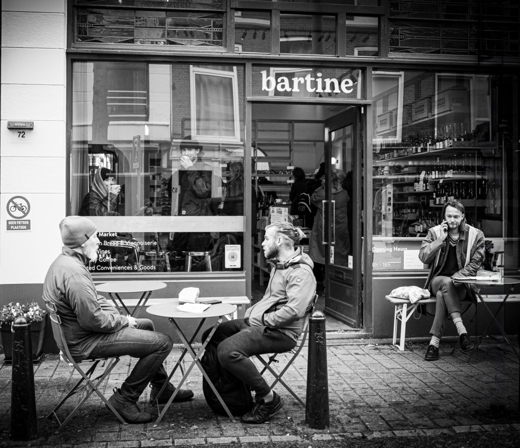 People outside Bartine enjoyin their coffee. Inside two people looking outside at the me and smiling. Black and white