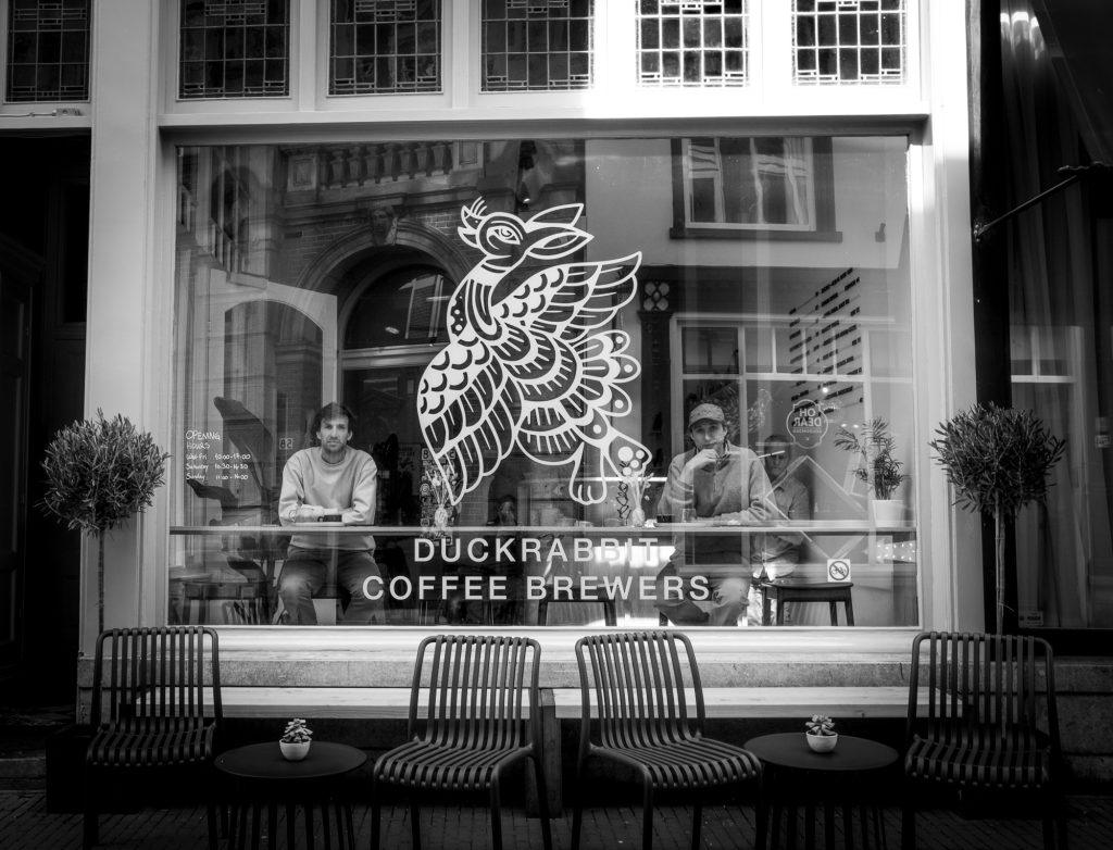 Front of DuckRabbit Coffee Brewers, tow pople sitting at the window looking ate me. Black and white